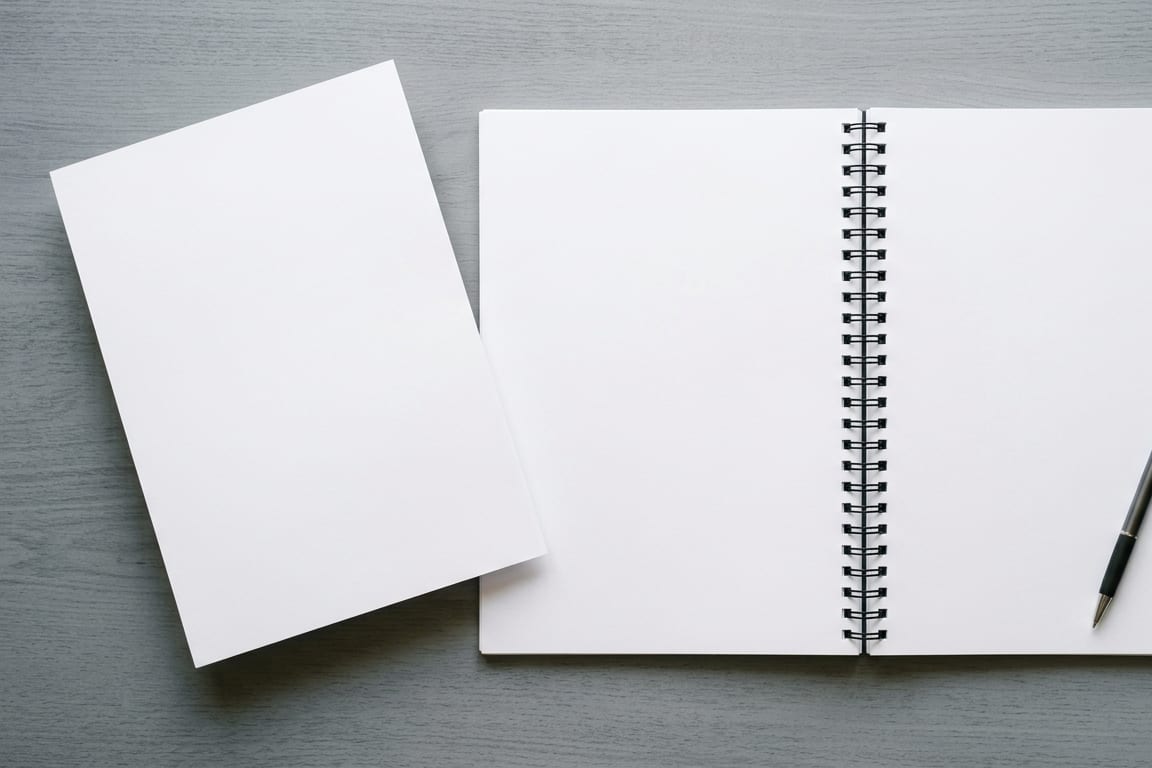An open blank notebook and a separate blank sheet laid side by side on a grey desk with a pen